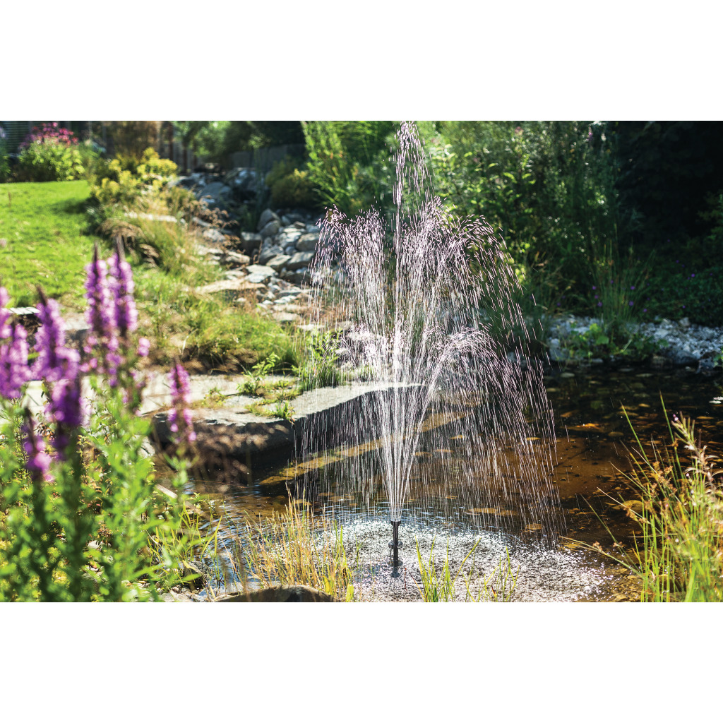 Une pompe de bassin Arosa projette une haute fontaine d'eau dans un bassin de jardin, entouré de végétation luxuriante et de fleurs violettes. À l'arrière-plan, un ruisseau rocailleux serpente au soleil. Des gouttes scintillent.