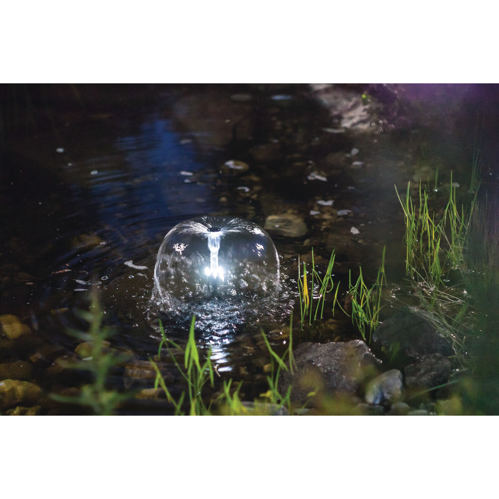 Un jet d'eau clair s'élève d'un étang sombre et forme une cloche d'eau en forme de dôme, éclairée par un anneau lumineux à LED. La lumière se reflète sur la surface légèrement ondulée. Des brins d'herbe et des pierres sur la rive.